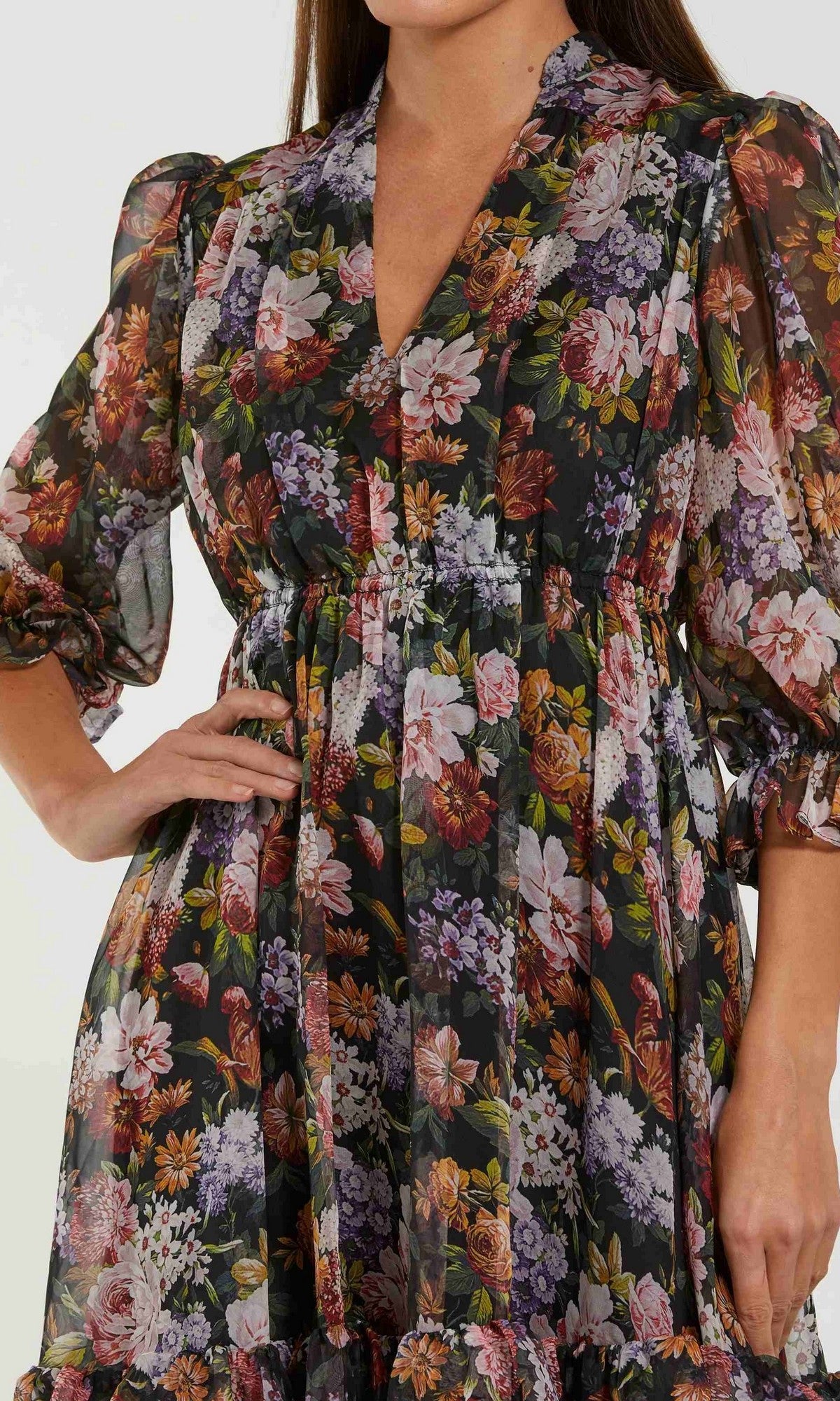 A woman wears the Mac Duggal 69031 Midi Floral Wedding-Guest Dress, featuring black chiffon with a floral pattern, long sheer sleeves, a gathered waist, and a V-neckline. Her left hand rests on her hip and only her upper body is visible.